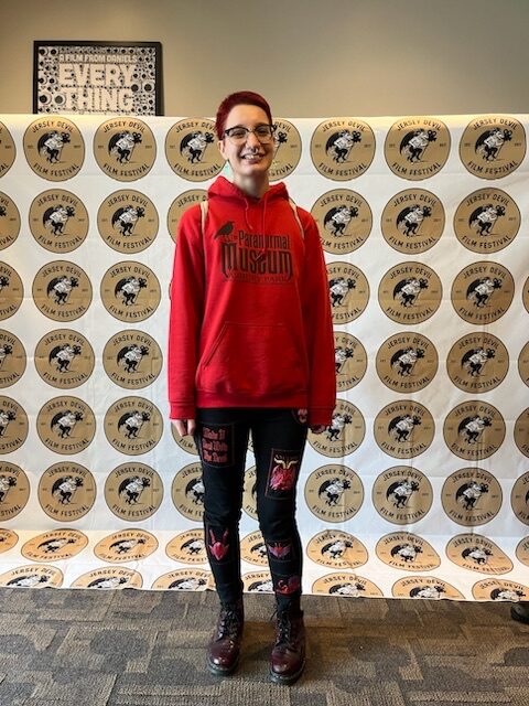 Woman in red hoodie and black pants standing in front of Jersey Devil Film Festival backdrop and merchandise.